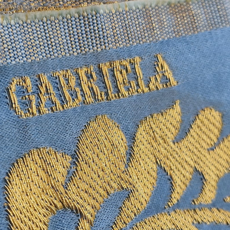May include: Close-up of a textile with the name "GABRIELA" embroidered in gold thread. The text is on a light blue fabric, with gold floral embroidery at the bottom. The top of the textile has a gold and white woven border.