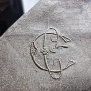 Puede incluir: Primer plano de una tela gris claro con un monograma bordado. El monograma presenta las letras "C" y "G" entrelazadas, con detalles florales y de hojas. El bordado es en un tono gris más claro.