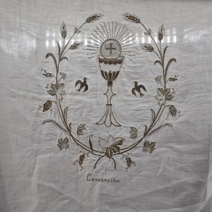 Puede incluir: Tela blanca con un diseño religioso bordado. El diseño presenta un cáliz, una cruz y un sol, rodeados de trigo y vides. También hay dos pájaros bordados. La palabra "Concepción" está escrita debajo del diseño.