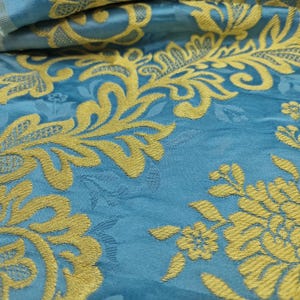 May include: Close-up of a teal fabric with a gold floral pattern. The design features stylized flowers and foliage, creating a textured, decorative surface. The fabric is folded, showing depth and detail in the weave.