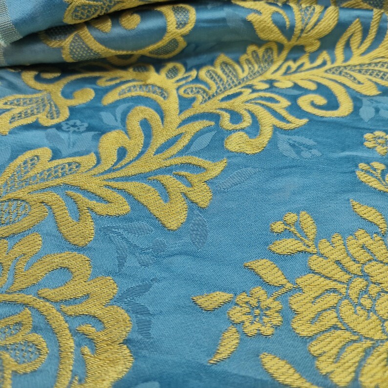 May include: Close-up of a teal fabric with a gold floral pattern. The design features stylized flowers and foliage, creating a textured, decorative surface. The fabric is folded, showing depth and detail in the weave.