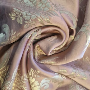 May include: Close-up of a luxurious, draped fabric in a soft rose color. The textile features an ornate, repeating floral pattern in a metallic gold hue. The fabric's folds and curves create a sense of depth and texture, highlighting its elegant design.