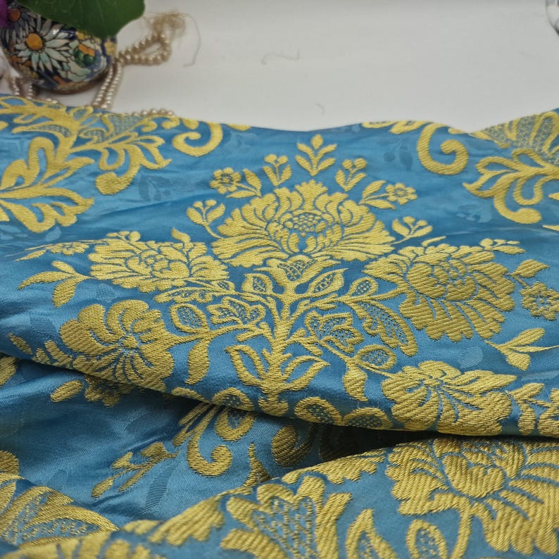 May include: Close-up of a blue fabric with a gold floral pattern. The fabric has a damask design with large, stylized flowers and leaves. The gold pattern stands out against the blue background, creating a luxurious appearance.