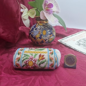 Puede incluir: Un pequeño estuche rectangular de cuentas con un patrón floral en rosa, rojo, verde y amarillo sobre un fondo crema con rayas verticales azules. Una moneda de un euro está colocada al lado para servir de escala y referencia de tamaño.