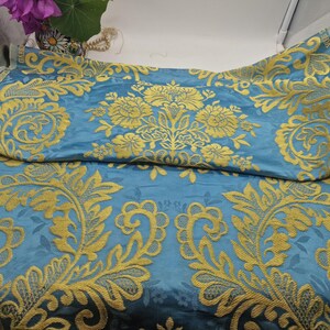 May include: Blue fabric featuring a gold floral damask pattern. The fabric has a silky sheen and is folded, showcasing the intricate design. The gold embroidery provides a striking contrast against the blue background. Suitable for upholstery or decorative projects.