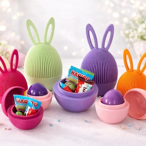 Könnte beinhalten: Eine Sammlung eiförmiger Behälter mit Hasenohren in Pink, Hellgrün, Lila und Orange. Einige Behälter sind geschlossen, während andere geöffnet sind und violette Kugeln sowie kleine Tüten mit Haribo-Süßigkeiten in den Basen zeigen.
