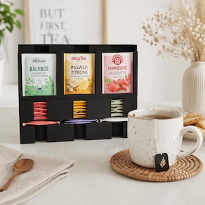 Könnte beinhalten: Ein schwarzer Teebeutel-Organizer mit drei oberen Fächern, die Teebeutel von Meßmer Balance, Yogi Tea Ingwer Zitrone und Pompadour Himbeere Hagebutte enthalten. Darunter befinden sich weitere Teebeutel in verschiedenen Farben. Eine gesprenkelte Keramiktasse steht auf einem runden geflochtenen Untersetzer.