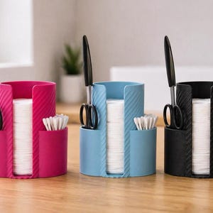 Könnte beinhalten: Drei zylindrische Badezimmer-Organizer in Pink, Hellblau und Schwarz. Jede Einheit verfügt über einen zentralen vertikalen Schlitz mit weißen Wattepads, ein Seitenfach mit weißen Wattestäbchen und einen Schlitz für eine schwarze Schere.
