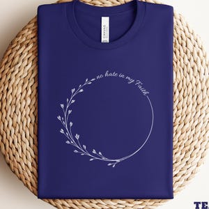 May include: Navy blue t-shirt with a white floral wreath design and the text "no hate in my Faith." The shirt is folded on a woven surface. The bottom right corner of the shirt has the text "TEAM NAVY."