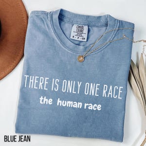 May include: Blue jean-colored t-shirt with the text "THERE IS ONLY ONE RACE the human race" in white. A gold necklace with a pendant is draped over the shirt. The shirt has a Comfort Colors tag.