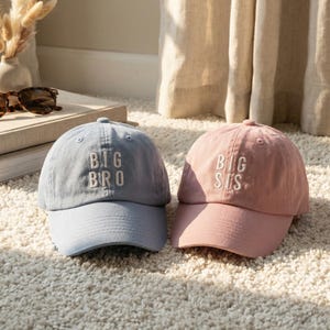 Puede incluir: Dos gorras de béisbol, una azul claro y otra rosa, sobre una alfombra blanca. La gorra azul claro tiene bordado en blanco "BIG BRO", mientras que la gorra rosa dice "BIG SIS". Las gorras son informales y adecuadas para el uso diario.