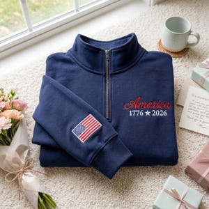 以下が含まれることがあります： ネイビーブルーのクォータージップスウェットシャツ。赤色の筆記体で「America 1776 2026」と刺繍されています。左袖にはアメリカ国旗のパッチが付いています。白いラグの上に折りたたまれています。