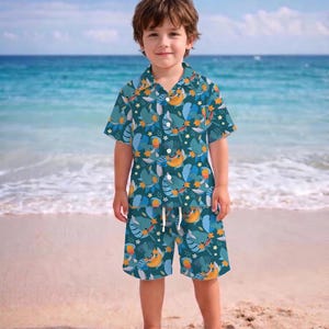 May include: A teal, short-sleeved button-down shirt and matching shorts set. The set features a tropical pattern with blue birds, yellow bananas, and green leaves. The shorts have a drawstring waist. The outfit is worn by a young child on a sandy beach.