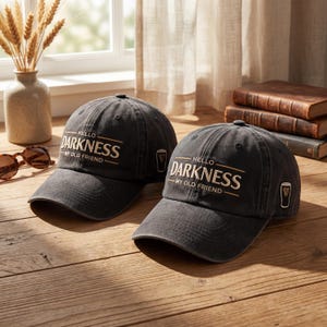 Op de afbeelding: Twee zwarte baseballpetten met de tekst "HELLO DARKNESS - MY OLD FRIEND" in gouden letters. Elke pet heeft een klein goudkleurig pintglaslogo. De petten liggen op een houten oppervlak.