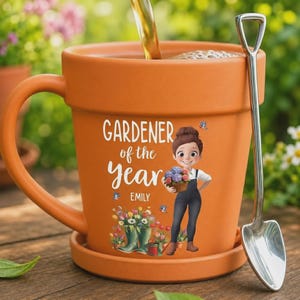 Puede incluir: Taza de cerámica naranja con forma de maceta, con un platillo y una cuchara a juego. La taza presenta el texto "GARDENER of the year" y una ilustración de dibujos animados de un jardinero. Se está vertiendo té en la taza.