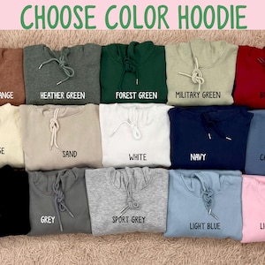 Op de afbeelding: Een selectie van opgevouwen hoodies in verschillende kleuren, waaronder heather orange, forest green en navy. De tekst "CHOOSE COLOR HOODIE" staat bovenaan. Elke hoodie heeft een trekkoord en is gelabeld met zijn kleurnaam.