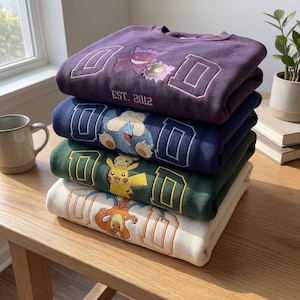 Puede incluir: Una pila de cuatro sudaderas en varios colores: morado, azul marino, verde y crema. Cada sudadera presenta un personaje Pokémon diferente y la letra "D" en un diseño de estilo universitario. La sudadera superior tiene la inscripción "EST. 2012".