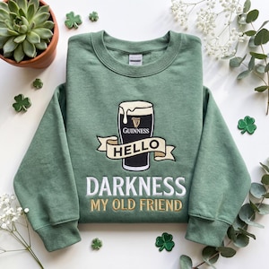 Op de afbeelding: Groen sweatshirt met een geborduurd Guinness pint-ontwerp en de tekst "Hello Darkness My Old Friend" in wit en goud. Klaverblad patches zijn op de achtergrond te zien.