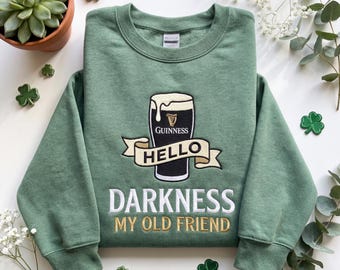 Op de afbeelding: Groen sweatshirt met een geborduurd Guinness pint-ontwerp en de tekst "Hello Darkness My Old Friend" in wit en goud. Klaverblad patches zijn op de achtergrond te zien.