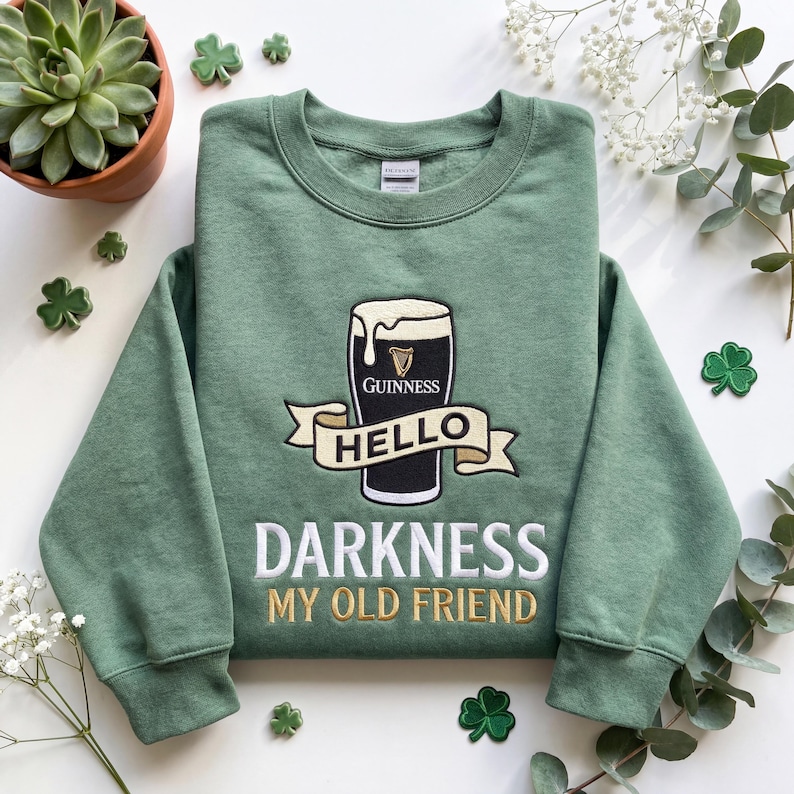 Op de afbeelding: Groen sweatshirt met een geborduurd Guinness pint-ontwerp en de tekst "Hello Darkness My Old Friend" in wit en goud. Klaverblad patches zijn op de achtergrond te zien.