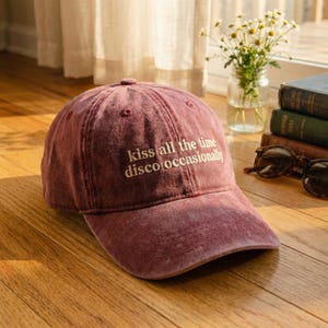 Könnte beinhalten: Weinrote Baseballkappe mit dem Text "kiss all the time disco occasionally" in Weiß gestickt. Die Kappe hat einen gebogenen Schirm und besteht aus einem weichen, abgenutzten Material. Der Hut liegt auf einer Holzoberfläche.