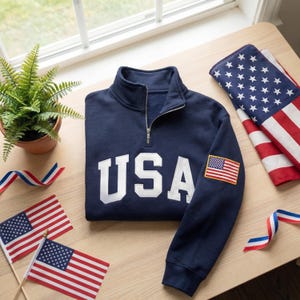 May include: A navy blue quarter-zip sweatshirt with the word "USA" in white. An American flag patch is on the left sleeve. The sweatshirt is on a wooden surface with small American flags and a folded American flag.