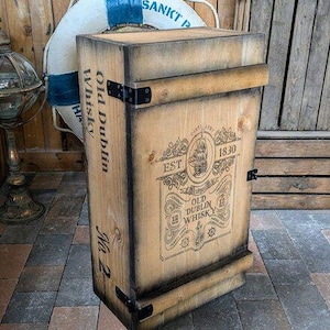 Può includere: Una cassa di legno con le parole "Old Dublin Whisky" e un'illustrazione di una nave. La cassa ha rinforzi angolari in metallo e un bordo tinto scuro. Un salvagente è dietro la cassa.