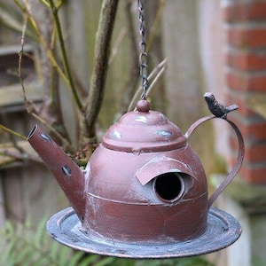 Könnte beinhalten: Ein Vogelhaus in Form einer Teekanne, braun lackiert mit einem Used-Look. Es hat einen kreisförmigen Eingang und einen kleinen Metallvogel am Griff. Hängt an einer Kette, perfekt für den Garten.