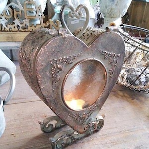 Op de afbeelding: Een hartvormige kandelaar in antieke stijl in zilver- en bronskleuren. De houder heeft sierlijke details, een helder glazen venster en een hartvormige handgreep. Hij staat op decoratieve voetjes en bevat een brandende kaars.