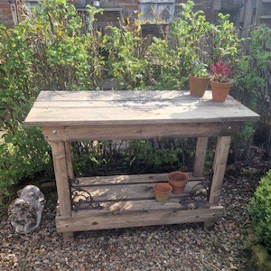 Peut inclure: Un établi de jardin en bois rustique avec une étagère inférieure et des supports métalliques décoratifs. Le banc en bois naturel accueille des pots de fleurs en terre cuite. Le fond présente de la verdure et un mur de briques.