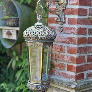 Può includere: Una lanterna da esterno in stile antico e ornato, con finitura bronzo. La lanterna presenta una parte superiore decorativa in filigrana e un involucro in vetro trasparente. È montata su un muro di mattoni con una staffa in bronzo abbinata. La lanterna è alta circa 46 cm.