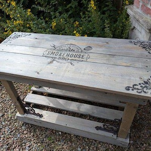 Op de afbeelding: Een grijze houten tafel met een onderste plank en decoratieve zwarte hoekaccenten. Het tafelblad heeft de tekst "SMOKEHOUSE BBQ" en een logo. De tafel wordt ondersteund door houten poten en is geschikt voor buitengebruik. Ongeveer 90 cm lang.