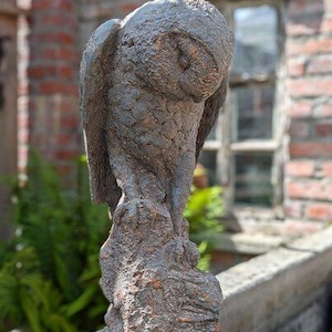 Könnte beinhalten: Eine detaillierte, verwitterte Statue einer Eule, die auf einem Felsen sitzt. Die Eule ist mit gesenktem Kopf dargestellt und zeigt strukturierte Federn und eine natürliche Farbpalette aus Braun- und Grautönen. Die Skulptur steht vor einem verschwommenen Hintergrund aus Ziegeln und Grün.