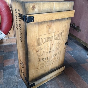 Può includere: Una cassa di legno con la scritta "Old Dublin Whisky Distillery" stampata sul davanti. La cassa è realizzata in legno marrone chiaro con cerniere e dettagli in metallo nero. La cassa ha un aspetto rustico e vintage.