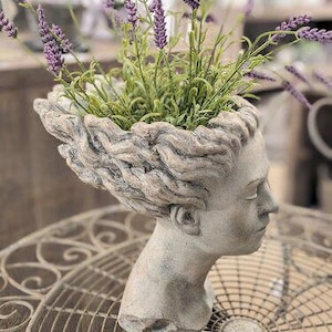 Può includere: Un vaso a forma di testa scolpita grigia con un volto sereno, riempito di lavanda finta. Il vaso ha un aspetto strutturato e invecchiato, e la lavanda presenta spighe di fiori viola e fogliame verde. Il vaso è su un tavolo di metallo decorativo.