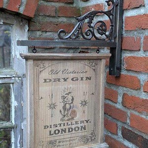 Op de afbeelding: Een houten bord voor "Old Victorian Dry Gin Distillery, London" met een decoratieve metalen beugel. Het bord is rechthoekig met een lichte houtnerf afwerking en heeft vintage letters en illustraties. De beugel is van donker metaal en gemonteerd op een bakstenen muur.