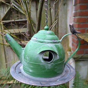 Può includere: Una casetta per uccelli in metallo verde a forma di teiera, appesa a una catena. Presenta un'apertura rotonda per gli uccelli e un piccolo ornamento a forma di uccello sul manico. La casetta è posizionata sopra un piattino di metallo.
