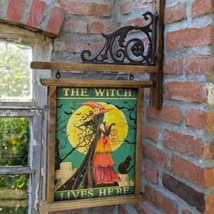 Könnte beinhalten: Holzschild mit dem Text "THE WITCH LIVES HERE" mit einer Hexenillustration vor einem Vollmond. Das Schild ist an einer dekorativen Metallhalterung und einem Holzrahmen befestigt, der an einer Ziegelmauer angebracht ist.