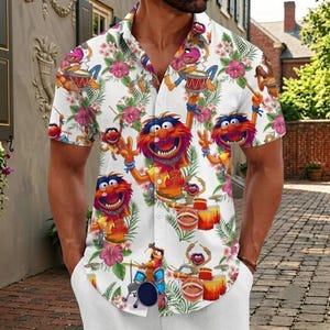 May include: A white short-sleeve button-down shirt featuring an all-over print of the Muppets character Animal playing drums, interspersed with pink hibiscus flowers and green tropical palm leaves. The shirt has a classic collar and front buttons.