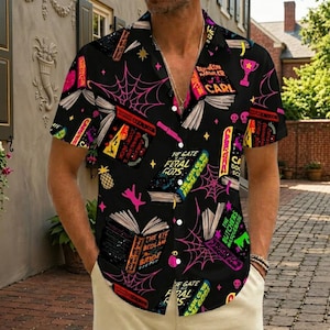 May include: A black short-sleeved button-down shirt featuring a repeating pattern of open books, pink spiderwebs, and small skulls. The book covers display titles such as The Gate of the Feral Gods, The Eye of the Bedlam Bride, and Dungeon Crawler Carl.