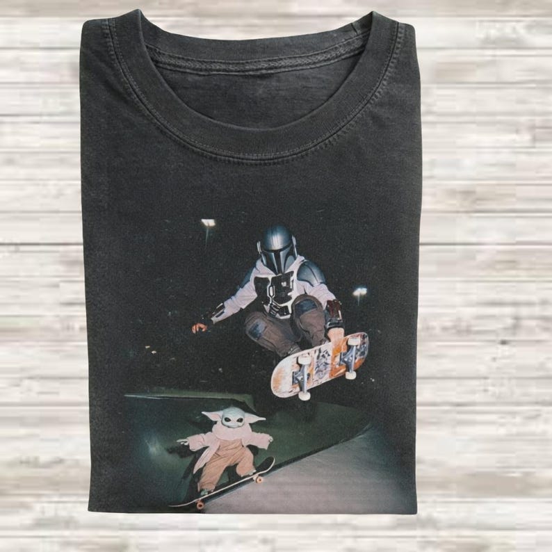 May include: A charcoal gray crew neck t-shirt featuring a graphic print of a character in a silver helmet and armor performing a skateboard trick in the air, while a small green creature in a tan robe stands on a skateboard below.