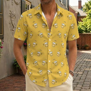 Op de afbeelding: Een shirt met korte mouwen en knoopsluiting in een levendige gele kleur, voorzien van een all-over herhalend patroon van het gezicht van Homer Simpson in zwarte lijnen. Het shirt heeft een klassieke kraag en een knoopsluiting aan de voorzijde.