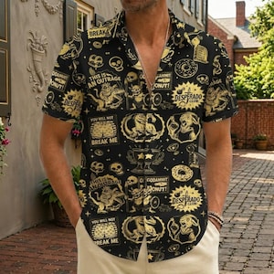 May include: A black short-sleeved button-down shirt featuring a repeating gold graphic print of skulls, trophies, doughnuts, and creatures. The text includes "THIS IS AN OUTRAGE", "YOU WILL NOT BREAK ME", "GODDAMMIT DONUT", and "DESPERADO CLUB" throughout the pattern.