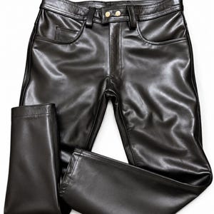 Könnte beinhalten: Schwarze Lederhose mit glatter, polierter Oberfläche, strukturiertem Bund mit Gürtelschlaufen, zwei Vordertaschen und einem vorderen Reißverschluss, der mit zwei goldfarbenen Metallknöpfen gesichert ist. Die Beine sind gerade und haben einen sauberen Saum.