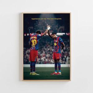 May include: Framed print featuring two soccer players in blue and red uniforms, high-fiving on a green field. The text "Superheroes are real. They wear blaugrana" is displayed at the top. The players are in a stadium setting.