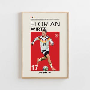 Puede incluir: Un póster enmarcado con el futbolista Florian Wirtz con una camiseta blanca, negra y dorada con el número 17. El póster tiene un fondo rojo con la palabra "GERMANY" en la parte inferior. El póster está en un marco de madera.