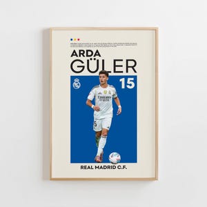 May include: A framed poster featuring soccer player Arda Güler in a white jersey with the number 15. The poster has a blue background with the Real Madrid C.F. logo and text. A soccer ball is at the player's feet.