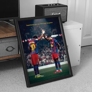 May include: Framed print featuring two soccer players in blue and red uniforms, high-fiving on a green field. The print includes the text "Superheroes are real. They wear blaugrana."