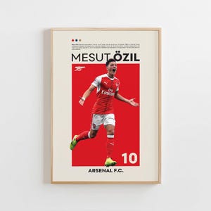 May include: A framed poster featuring Mesut Özil in an Arsenal F.C. uniform. The poster has a red background with the player's image, the number 10, and the team's name. The poster includes text at the top and bottom.