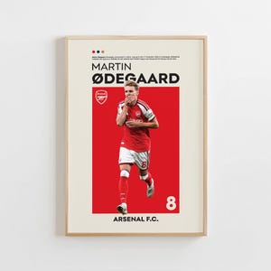 May include: A framed poster featuring a soccer player, Martin Ødegaard, in a red and white Arsenal F.C. kit. The design includes the player's name, the Arsenal crest, the number 8, and text detailing his professional background on a cream background.